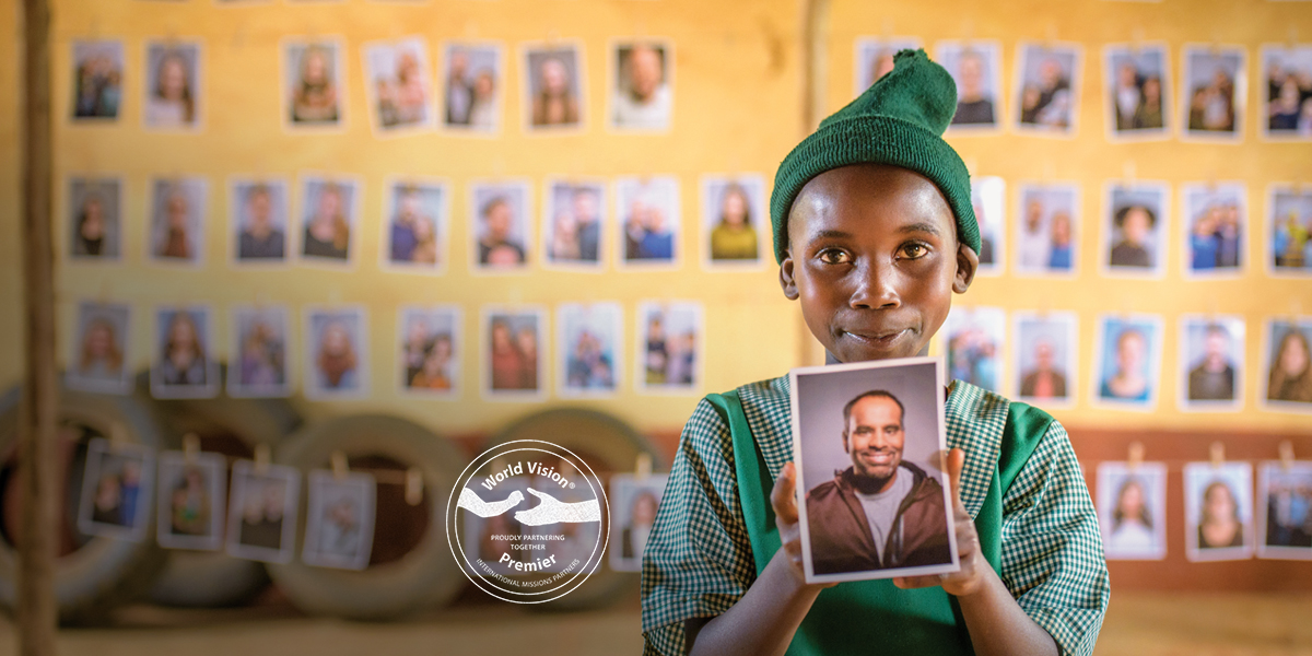 Rosemary from Kenya smiles and holds up a photo of the sponsor she has chosen. There are rows of photos of other sponsors in the background behind her