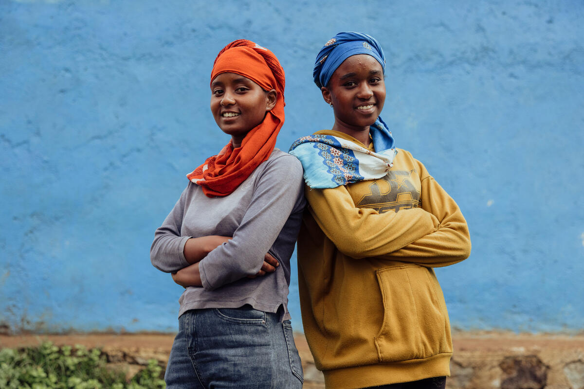 Two teenage girls stand back to back, arms crossed, smiling to camera