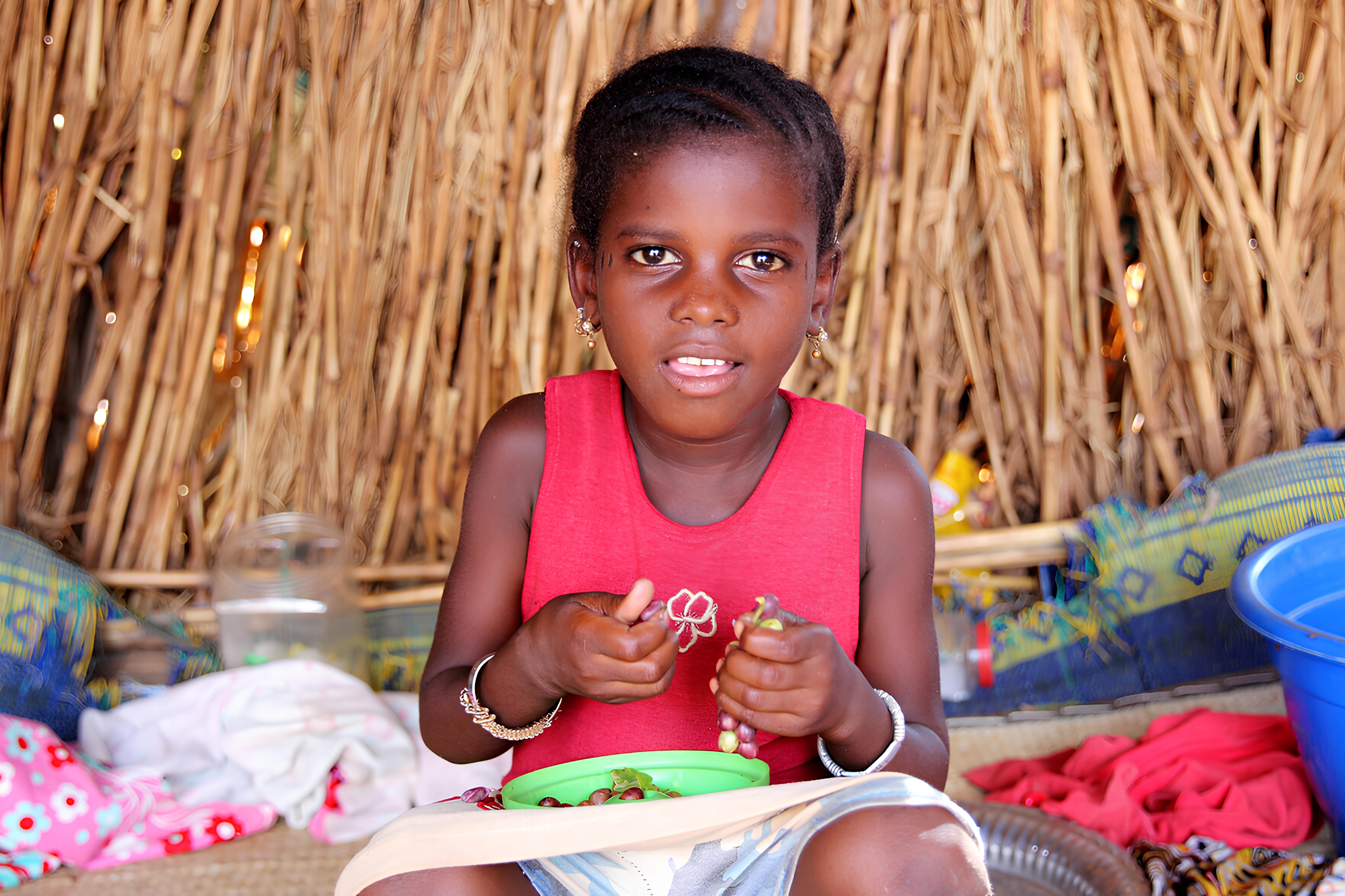 Young girl sitting in front of a thatched backdrop, eating some grapes and looking into the camera