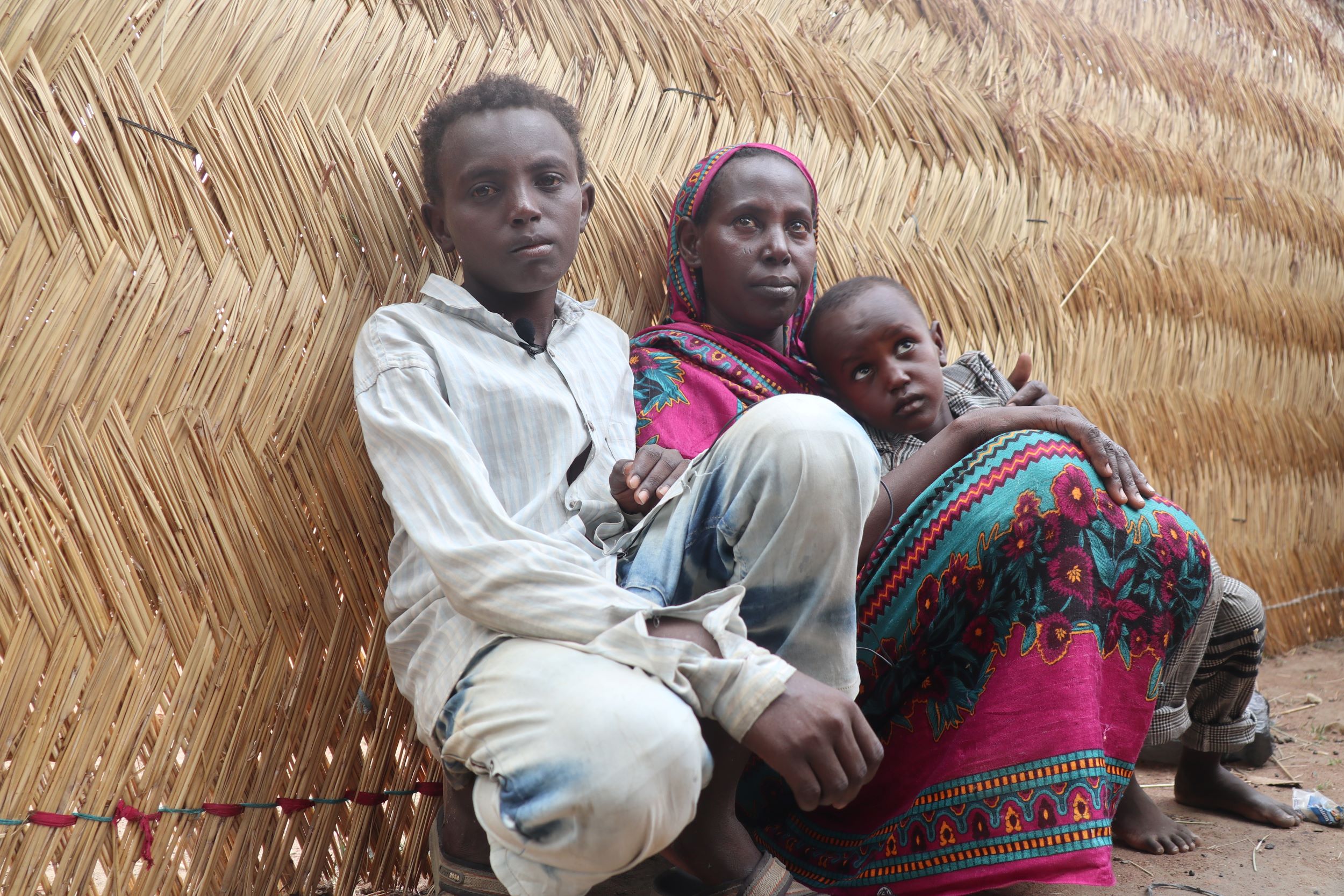 A refugee boy sits with his mum and sibling outside against the woven wall of their shelter, South Sudan