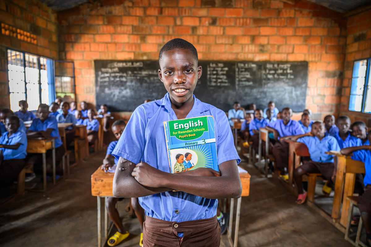 Amideu, 17, stands out the front of a classroom in Rwanda holding his school books, with classmates sitting in rows behind him.