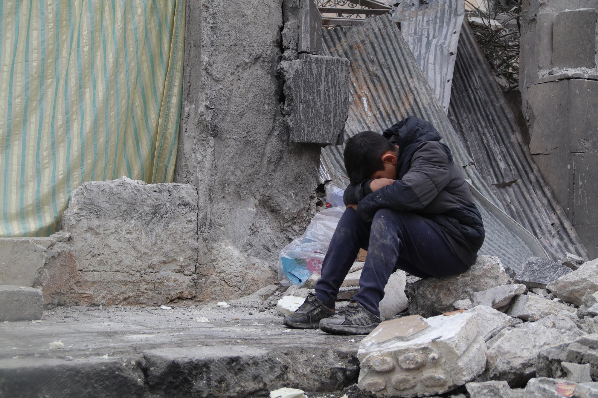 A boy sits in front of destroyed buildings with his head in his hands