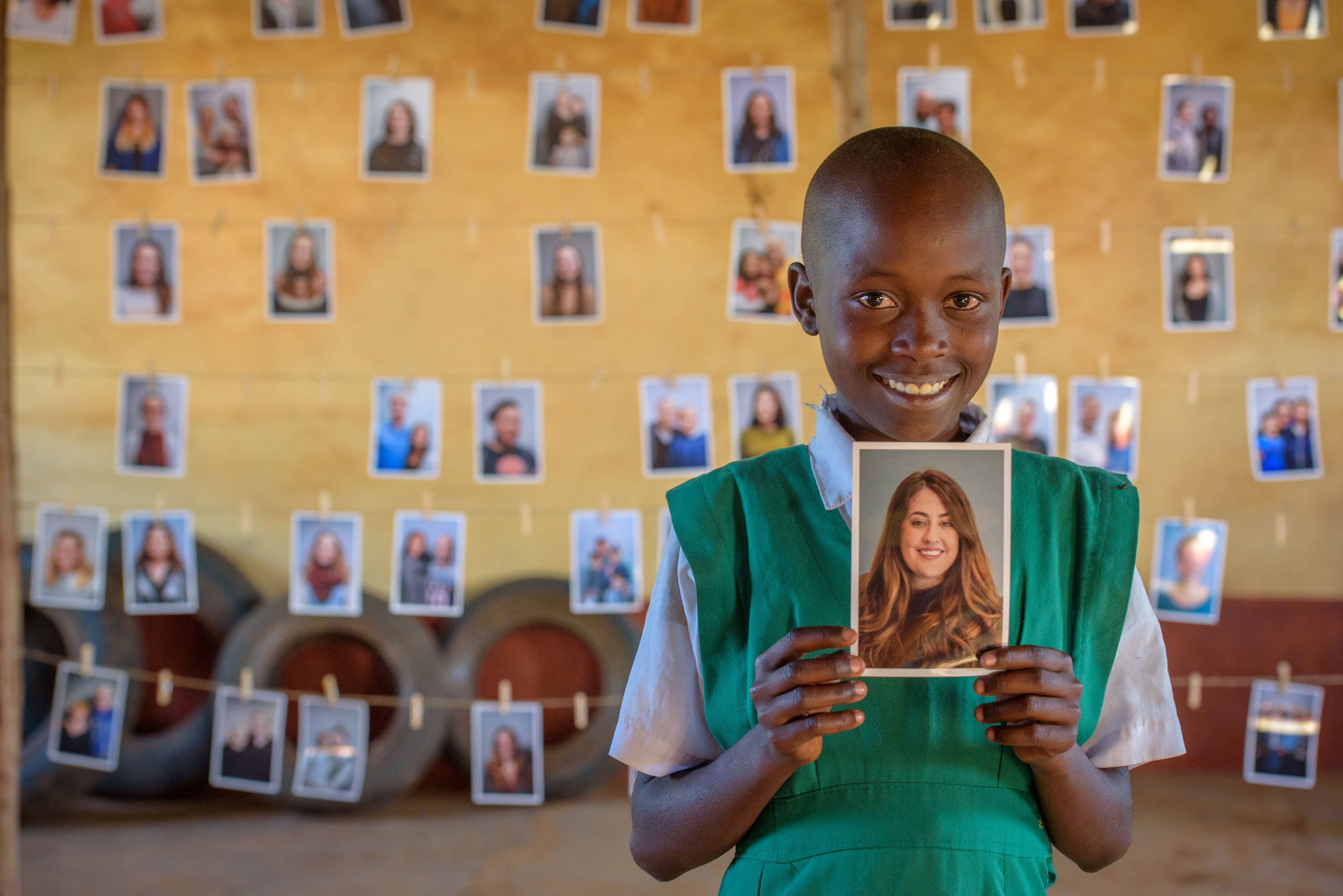 Anna from Kenya smiles and holds up a photo of the sponsor she has chosen. There are rows of photos of other sponsors in the background behind her