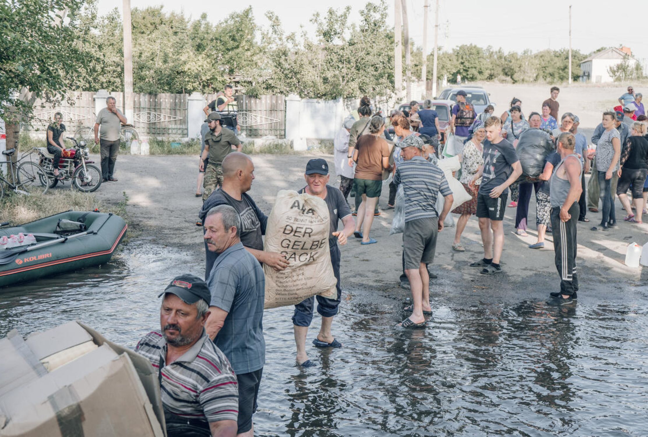 A line of Ukrainian people wading through floodwaters carrying bags containing food and hygiene kits.